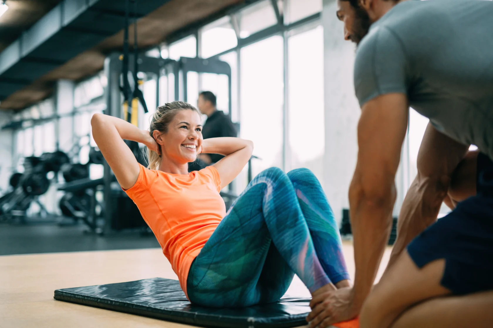 A woman in an orange shirt performs sit-ups on a mat while a man assists her in a gym setting.