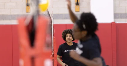 Two people playing volleyball indoors, one person in focus watching the game while the other is blurred in action with a raised arm.