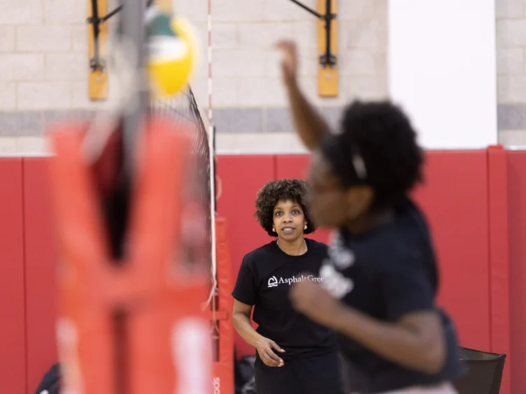 Two people playing volleyball indoors, one person in focus watching the game while the other is blurred in action with a raised arm.