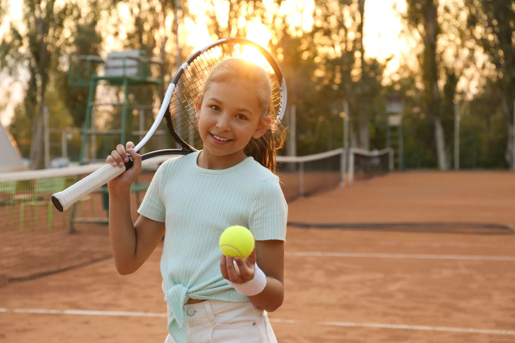 Young girl in a light blue shirt holding a tennis racket and ball on a clay tennis court with trees and a bright sky in the background.