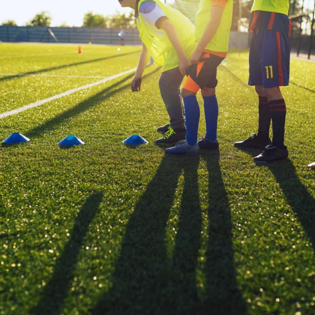 Children in sports uniforms line up for soccer drills on a grassy field near blue cones. Shadows are cast on the grass.