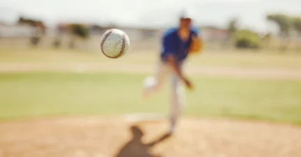 A baseball is mid-air with a pitcher in a blue jersey throwing it from the mound on a sunny day at the field.