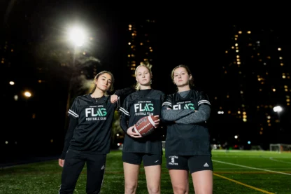 Three people in matching uniforms stand on a lit sports field at night, holding a football, with city lights in the background.