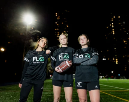 Three people in matching uniforms stand on a lit sports field at night, holding a football, with city lights in the background.