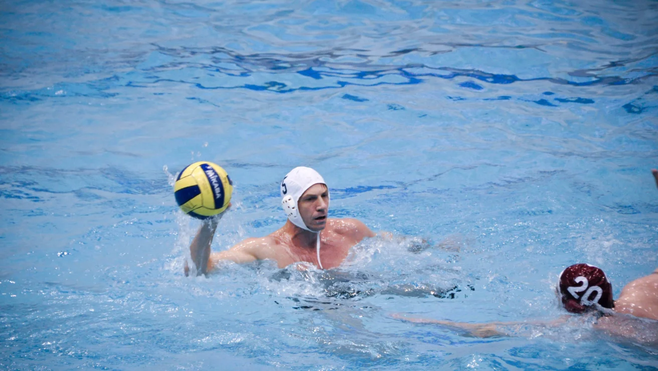 Two water polo players in a pool, one wearing a white cap holding a ball above the water, while the other in a red cap tries to block.