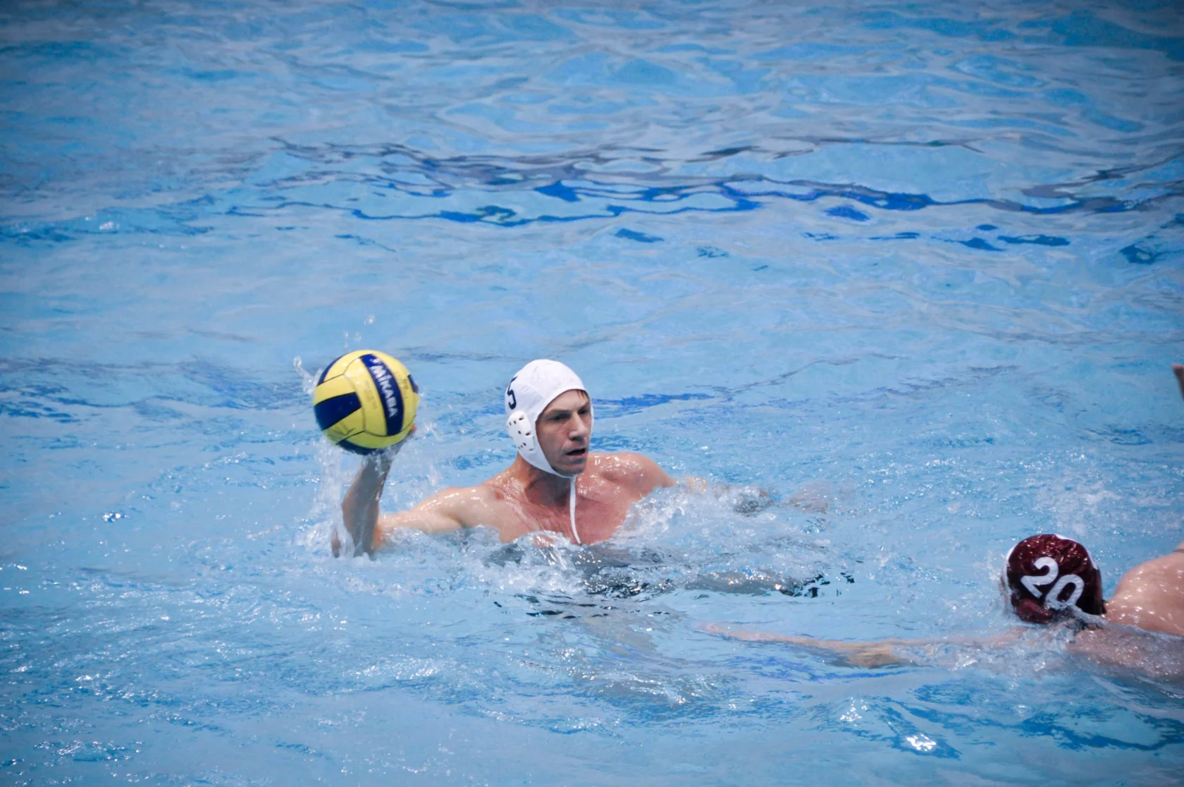 Two water polo players in a pool, one wearing a white cap holding a ball above the water, while the other in a red cap tries to block.