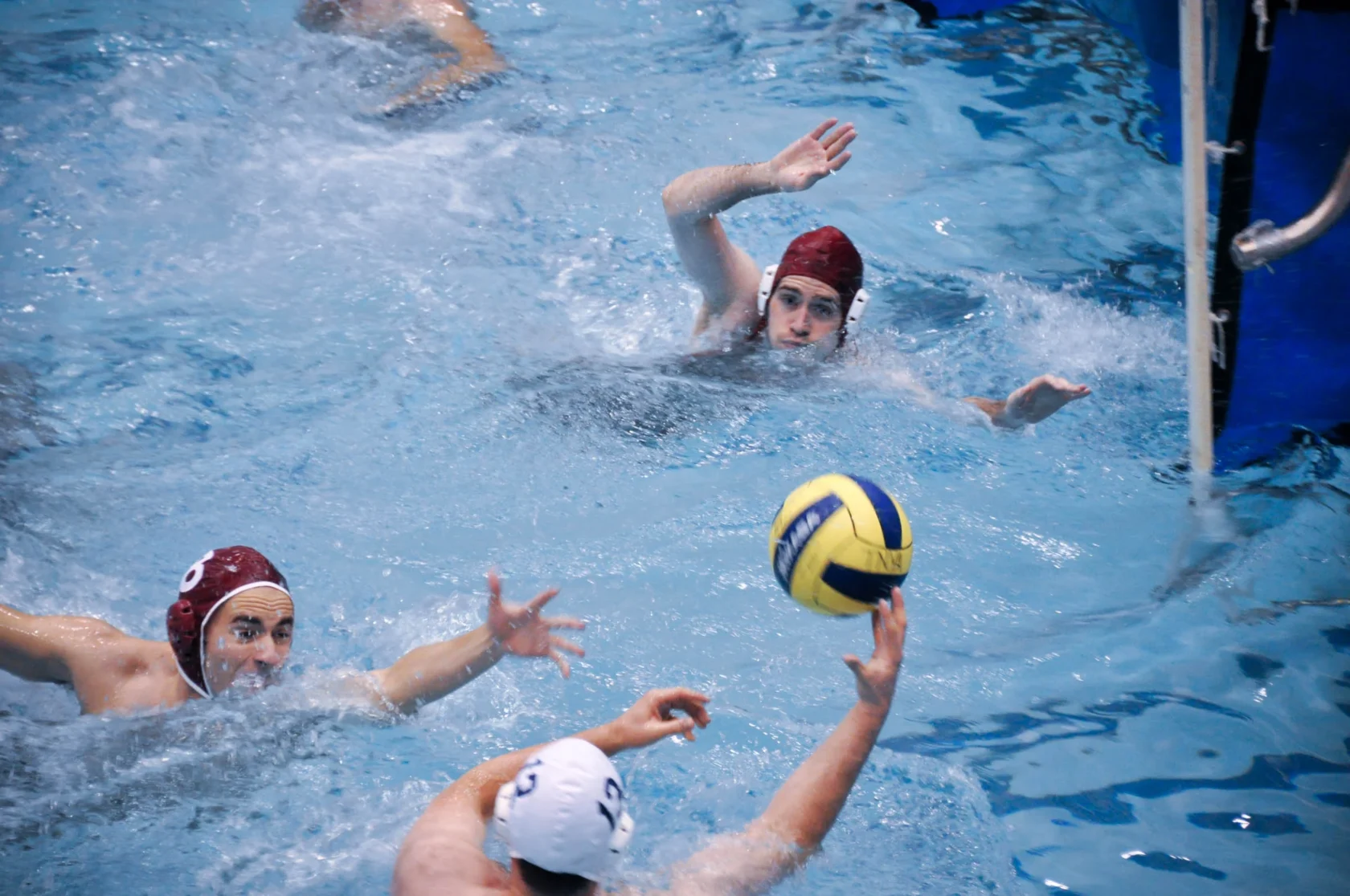 Water polo players in a pool, with one player reaching for the ball and others nearby, all wearing caps.