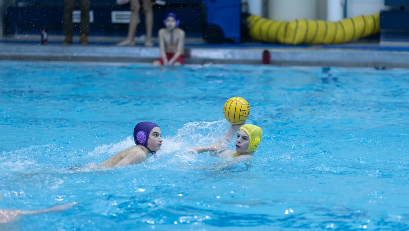 Two water polo players in action. One player in a purple cap faces an opponent in a yellow cap holding the ball above water. A third person observes from the poolside in the background.