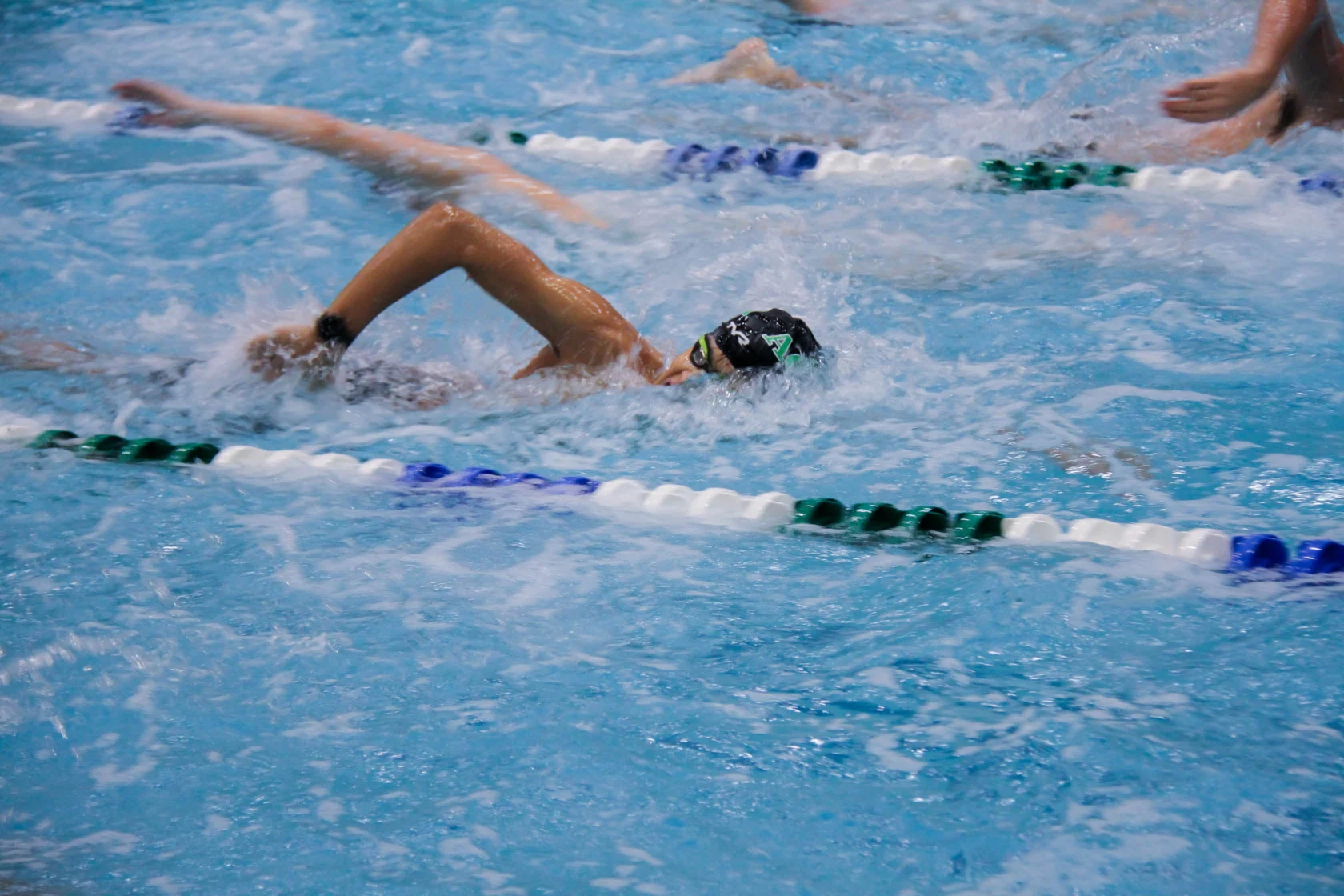 A swimmer in a black cap competes in a pool, surrounded by other swimmers, separated by lane dividers.