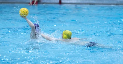 Two water polo players in a pool compete for the ball; one player wears a yellow cap and the other a purple cap.