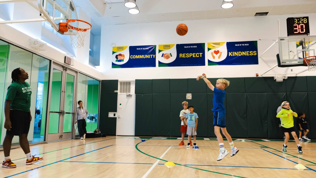 Children play basketball in a gym, practicing shooting hoops, while an instructor observes. Banners on the wall display "Community," "Respect," and "Kindness.