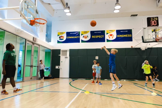 Children play basketball in a gym, practicing shooting hoops, while an instructor observes. Banners on the wall display "Community," "Respect," and "Kindness.