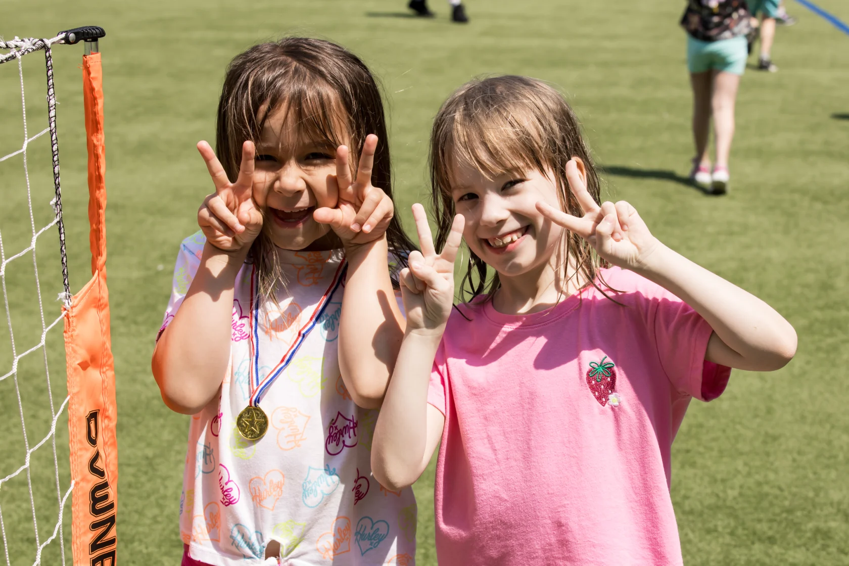 Two young girls smiling and making peace signs next to a soccer goal on a grassy field. One girl wears a medal.