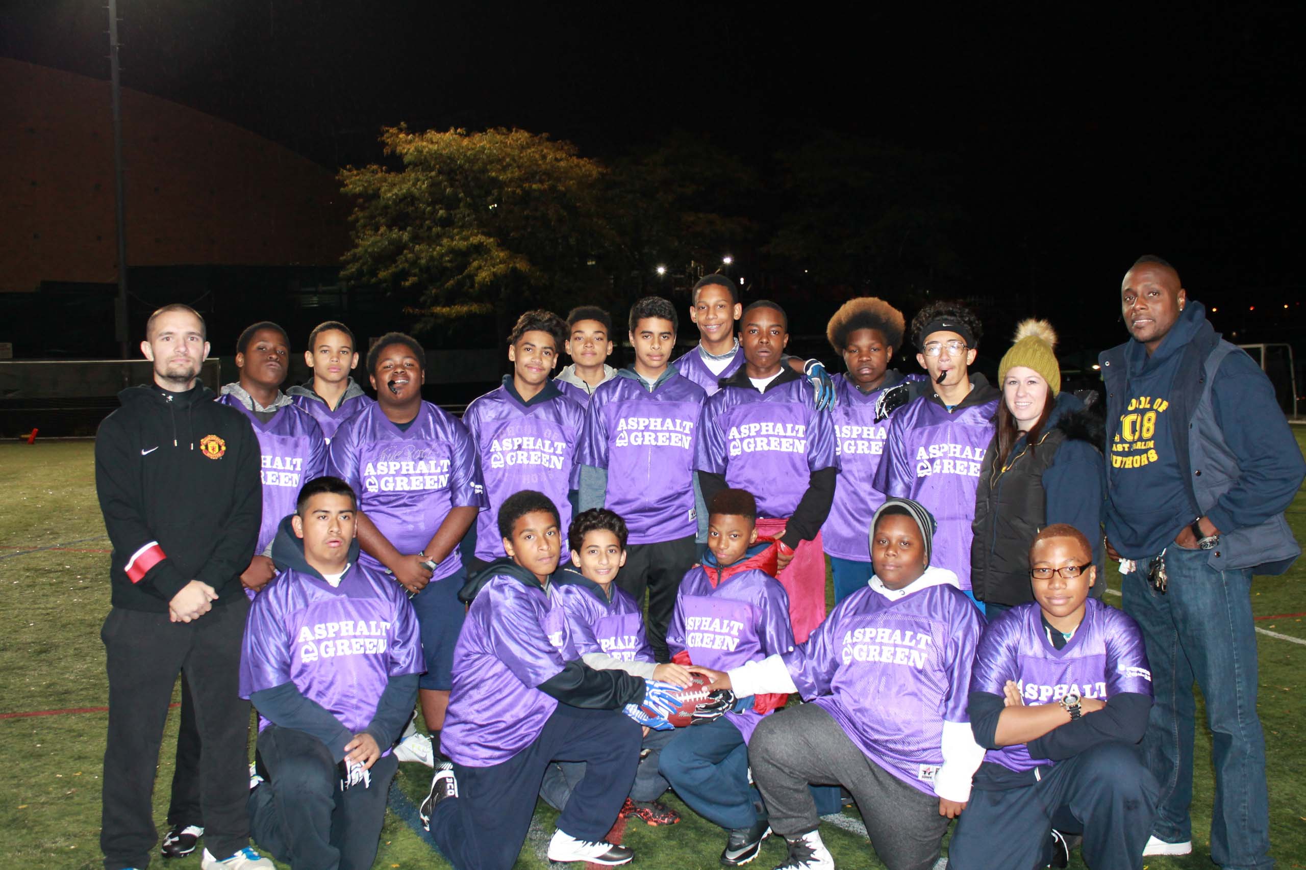 A group of people wearing matching purple jerseys poses for a photo on a field at night.
