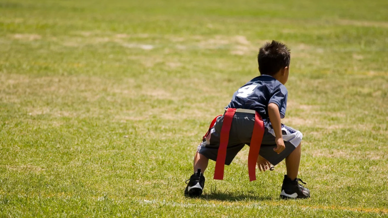 Child in a football uniform crouches on a grassy field, wearing a jersey with the number 4 and a flag attached to his waist.