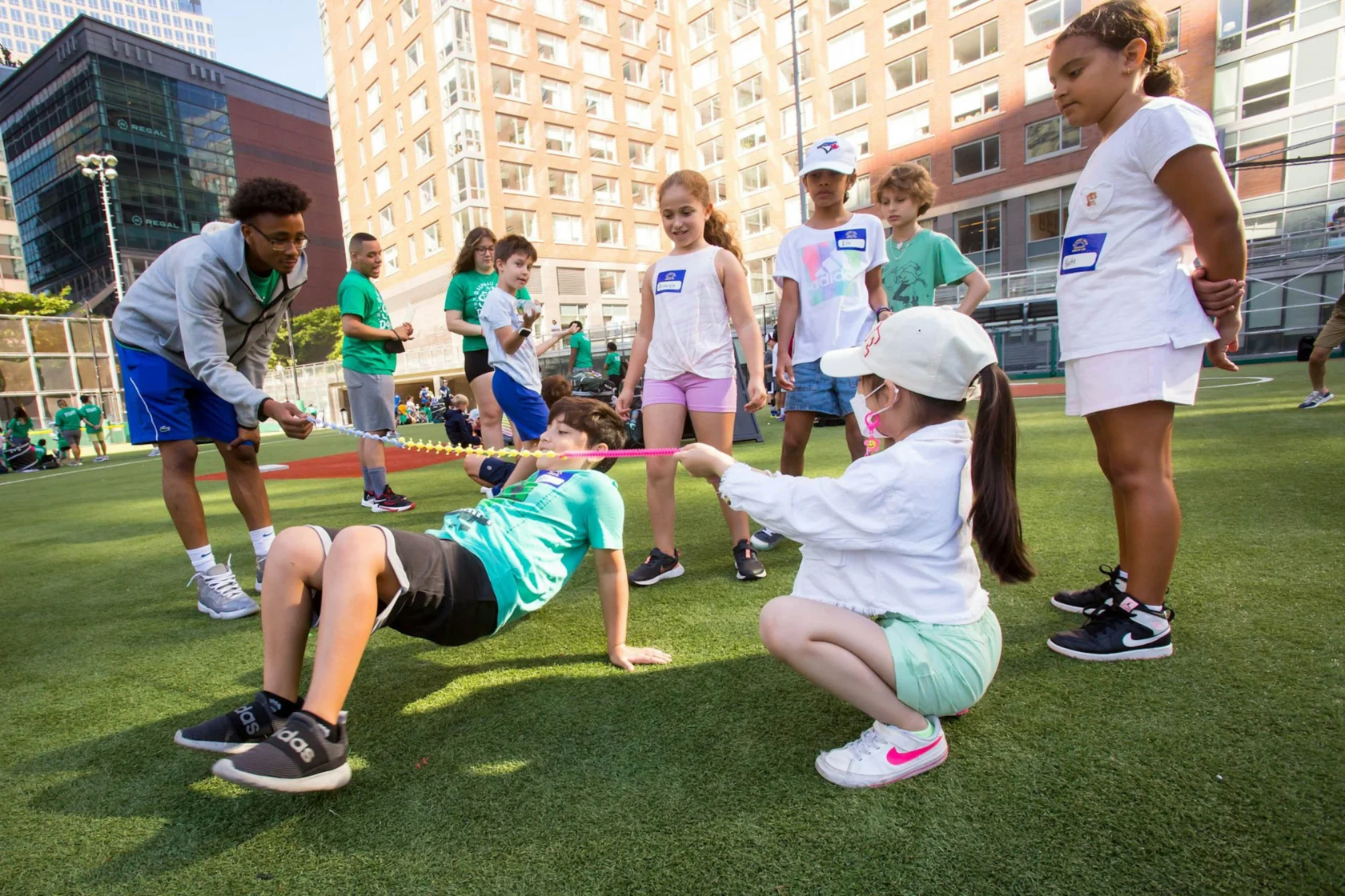 A group of children and an adult participate in an outdoor limbo game on a grass field. Buildings are visible in the background.