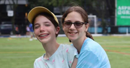 Two girls smiling outdoors, one wearing a yellow cap and braces, the other with glasses, in a park setting.