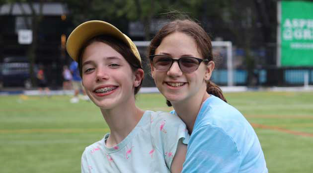 Two girls smiling outdoors, one wearing a yellow cap and braces, the other with glasses, in a park setting.