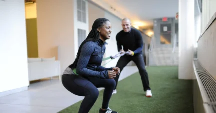 A woman performs squats with a resistance band, assisted by a man in a hallway with artificial turf flooring.