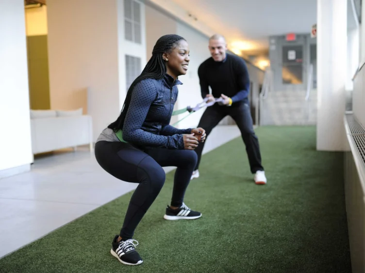 A woman performs squats with a resistance band, assisted by a man in a hallway with artificial turf flooring.