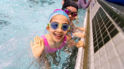 A smiling child wearing goggles and a swim cap waves from a pool, with another person partially visible in the background.
