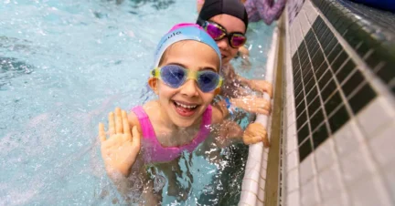 A smiling child wearing goggles and a swim cap waves from a pool, with another person partially visible in the background.