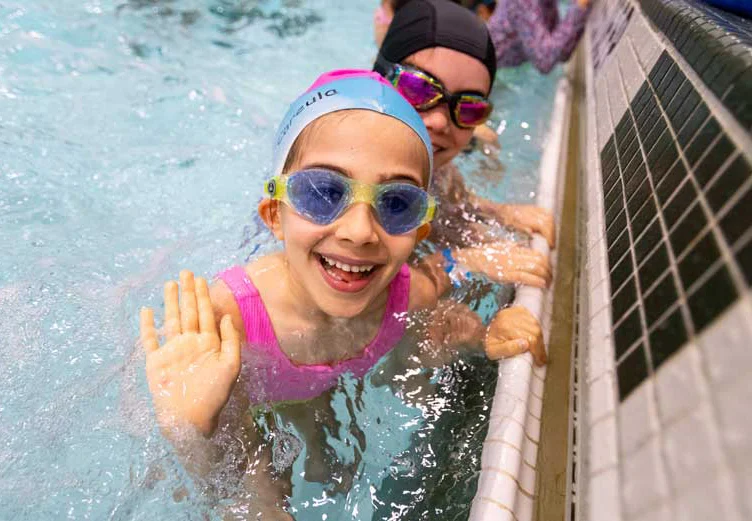 A smiling child wearing goggles and a swim cap waves from a pool, with another person partially visible in the background.