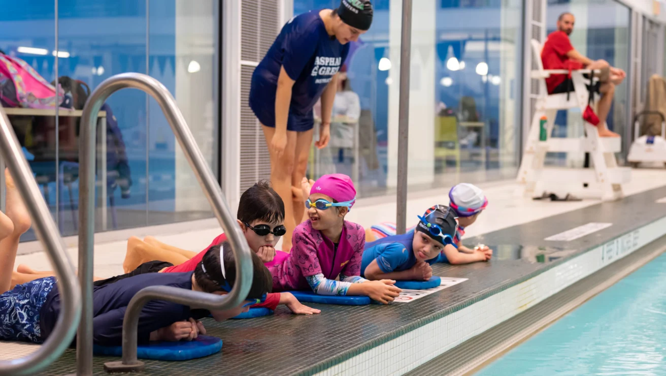 Children wearing swim gear lie on the poolside while an instructor supervises. A lifeguard sits nearby.