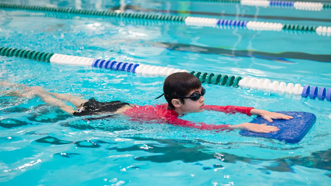 A child wearing goggles and a red shirt uses a kickboard while swimming in a pool lane.