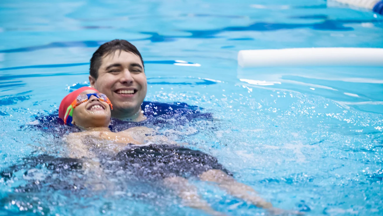 A child wearing goggles and a swim cap is floating on their back in a swimming pool, supported by an adult who is smiling.