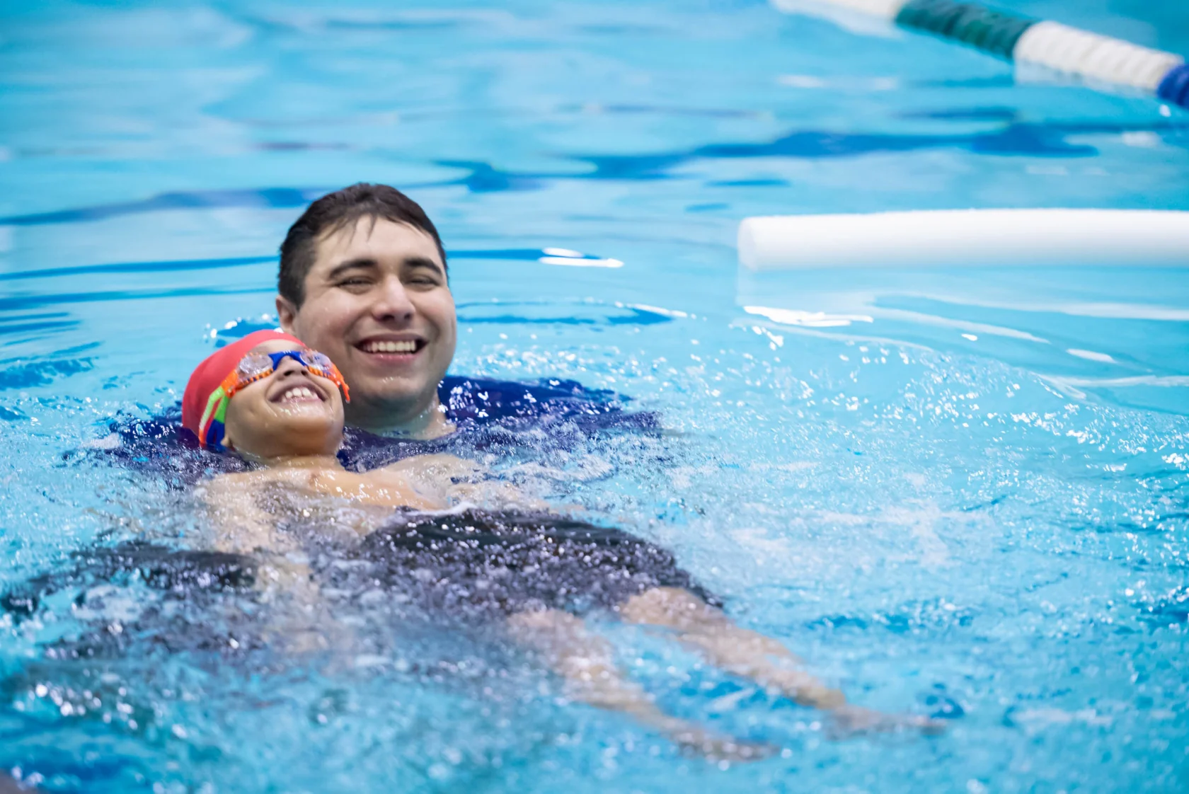 A child wearing goggles and a swim cap is floating on their back in a swimming pool, supported by an adult who is smiling.