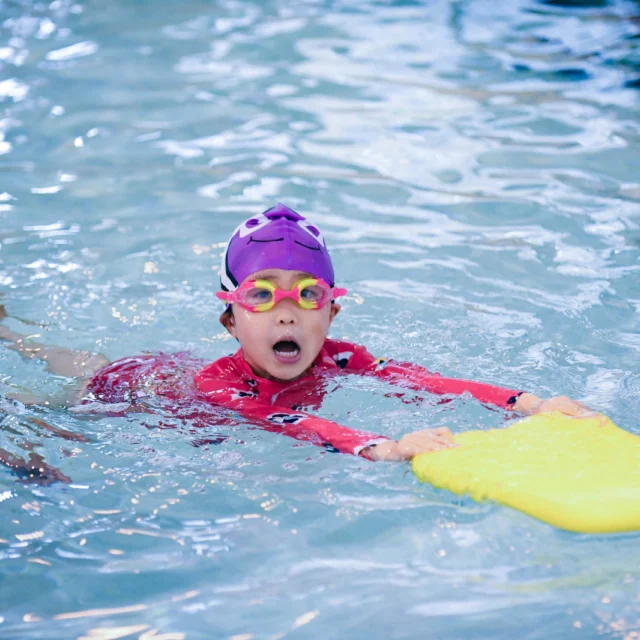 Child in a purple cap and goggles practices swimming with a yellow kickboard in a pool. An adult wearing a swim cap is nearby.