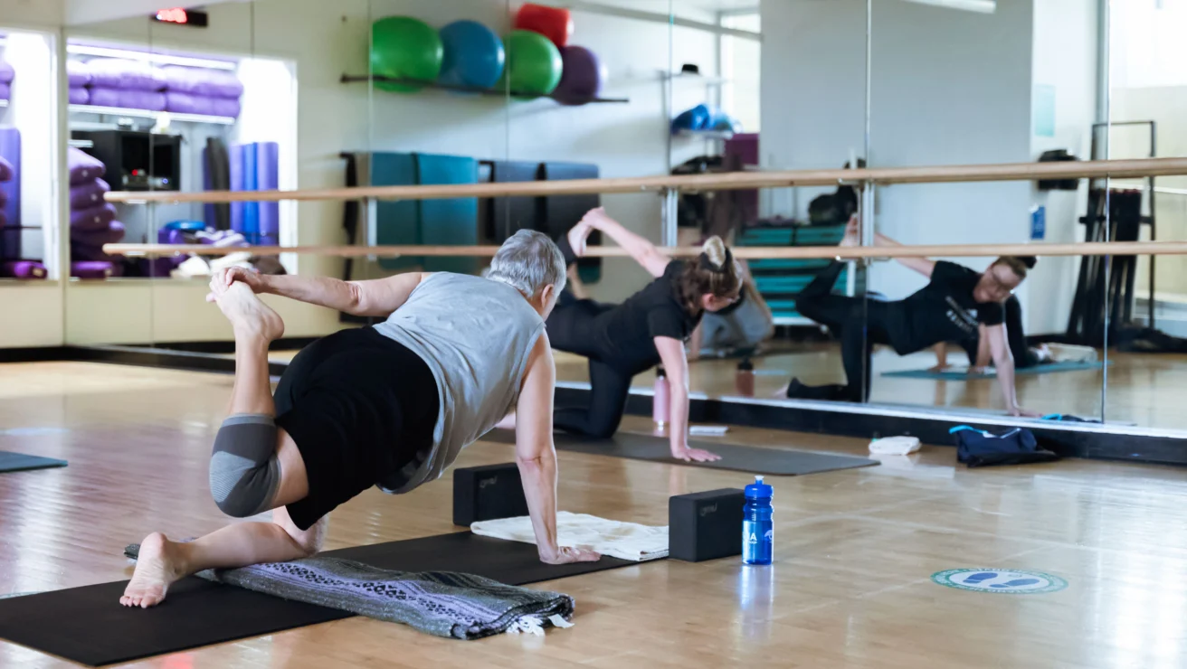 Two people practicing yoga in a studio, performing floor stretches in front of mirrors, with yoga mats and a water bottle visible.