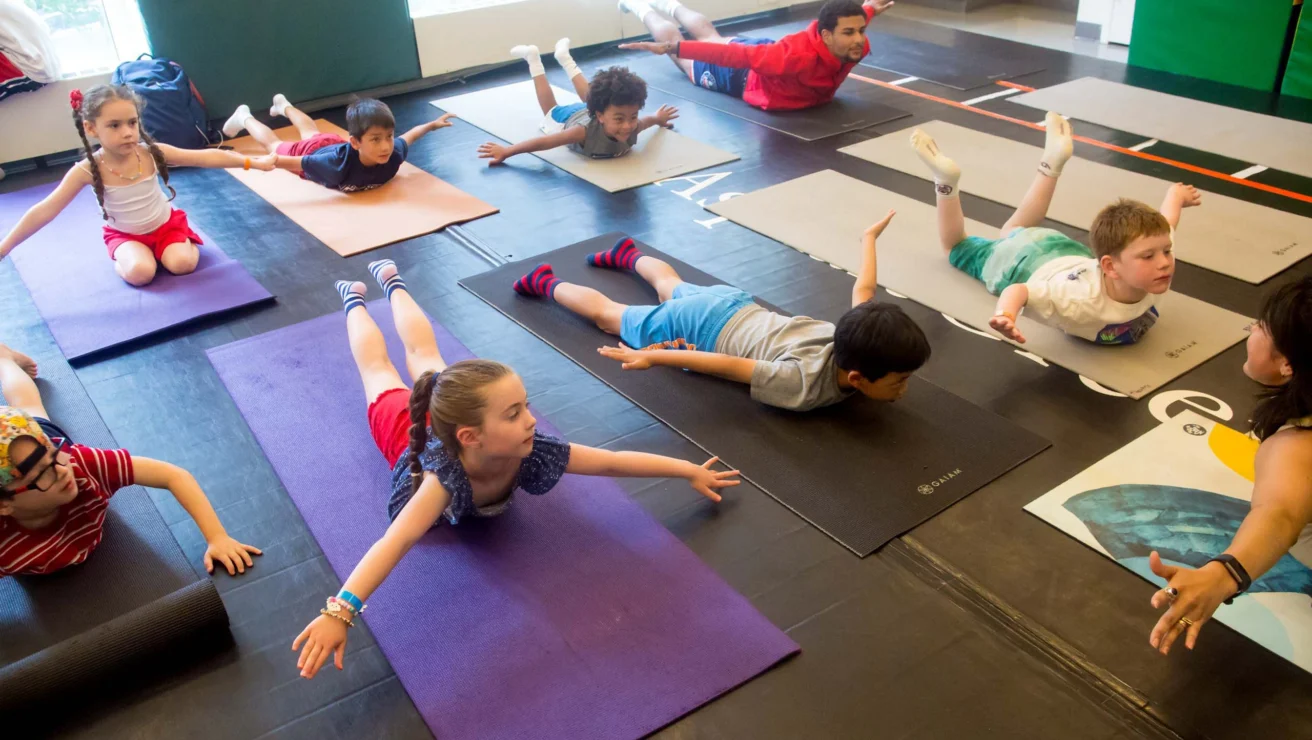 Children are lying on yoga mats in a classroom, performing a pose with their arms and legs lifted off the ground, guided by a teacher.