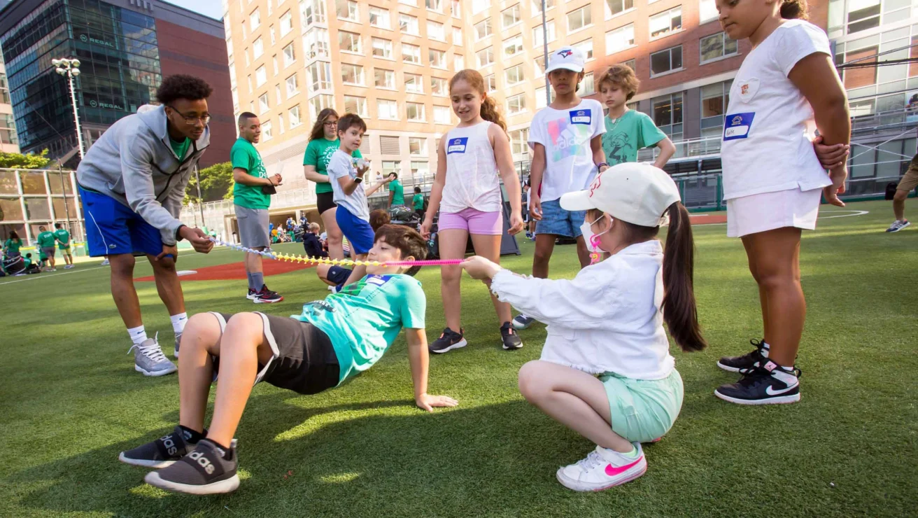 Children participate in an outdoor game on a sports field, with a coach assisting and others watching.