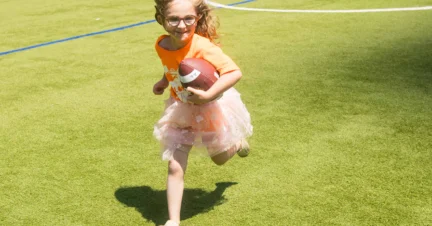 A girl in an orange shirt and skirt runs on a grassy field while holding a football.