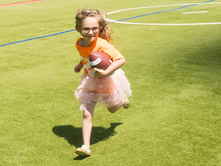 A girl in an orange shirt and skirt runs on a grassy field while holding a football.
