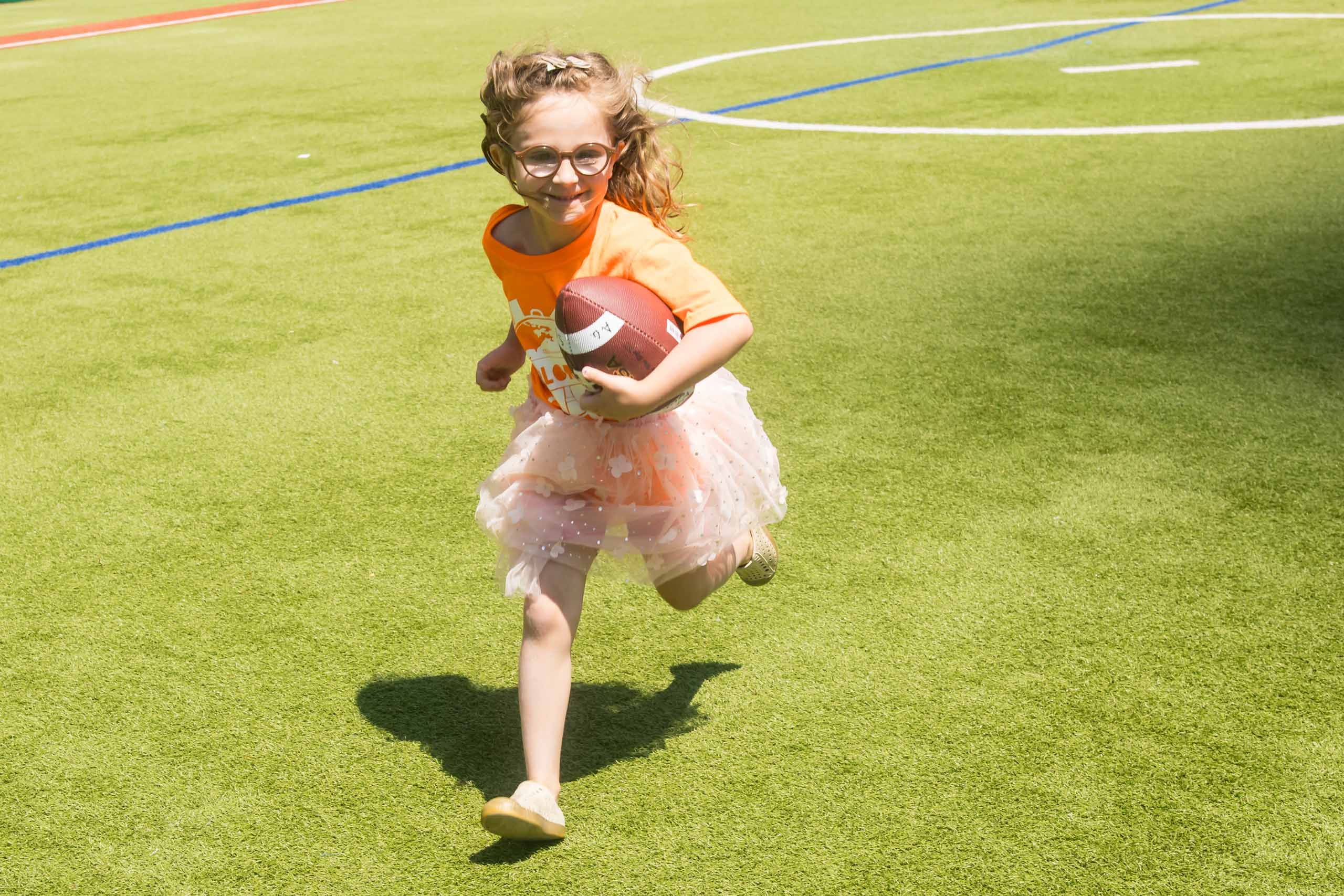 A girl in an orange shirt and skirt runs on a grassy field while holding a football.