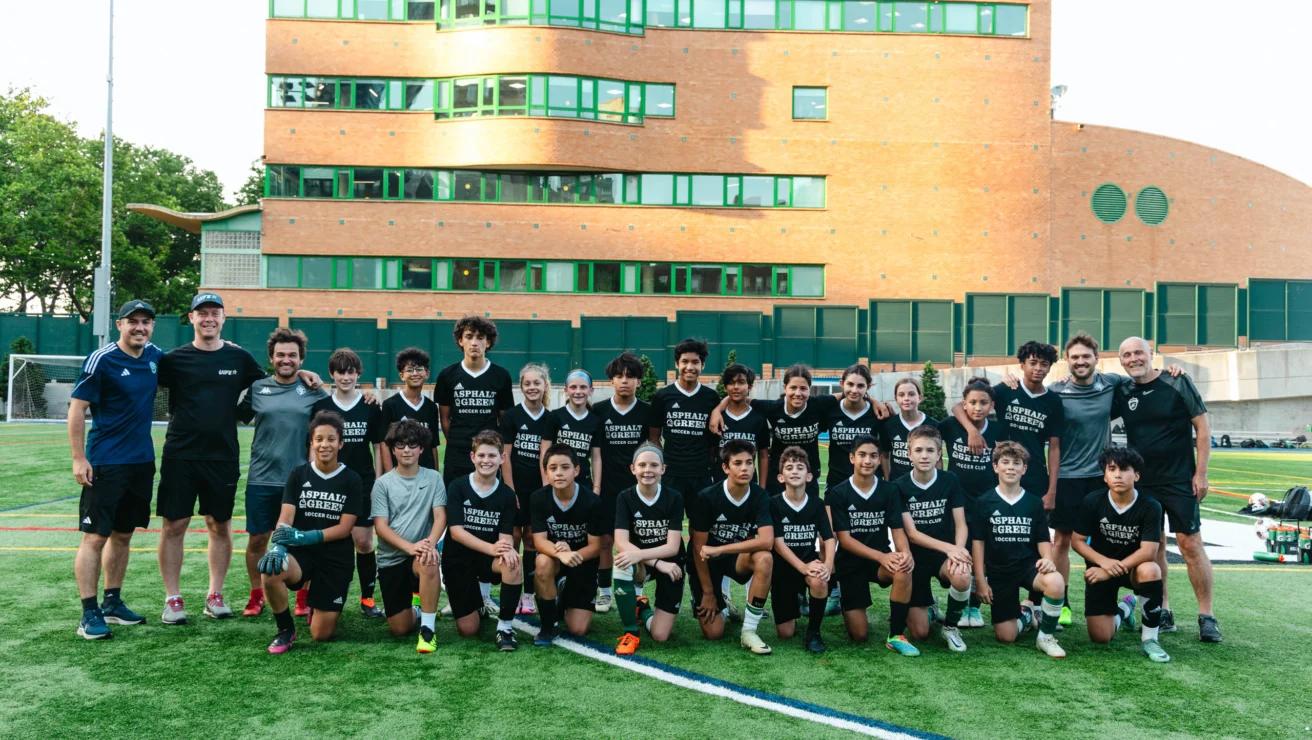 A soccer team poses for a group photo on a field, with coaches standing on either side. A brick building is in the background.