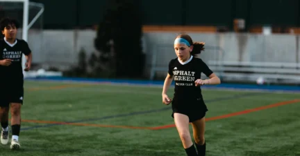 A young soccer player in black kit dribbles the ball on a field, with a teammate in the background.