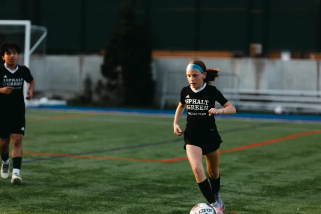 A young soccer player in black kit dribbles the ball on a field, with a teammate in the background.