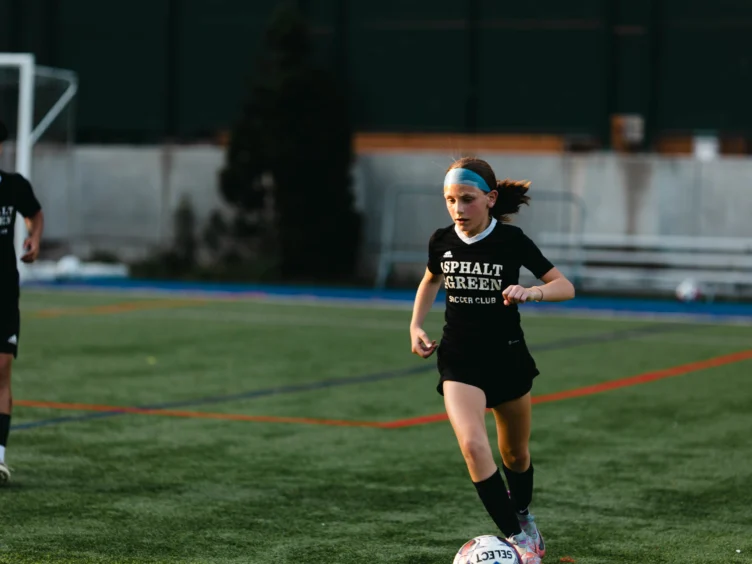 A young soccer player in black kit dribbles the ball on a field, with a teammate in the background.
