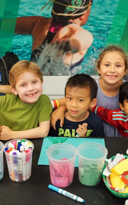 Five children smiling behind a table with arts and crafts supplies, including markers, foam shapes, and colored paper.