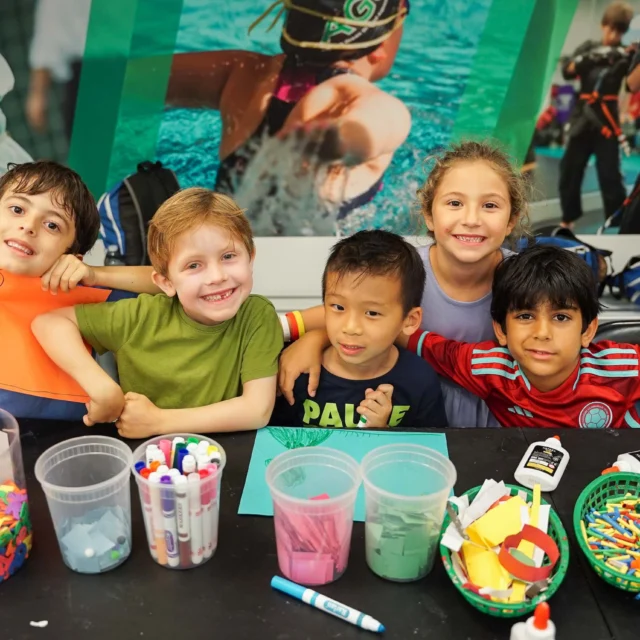 Five children smiling behind a table with arts and crafts supplies, including markers, foam shapes, and colored paper.