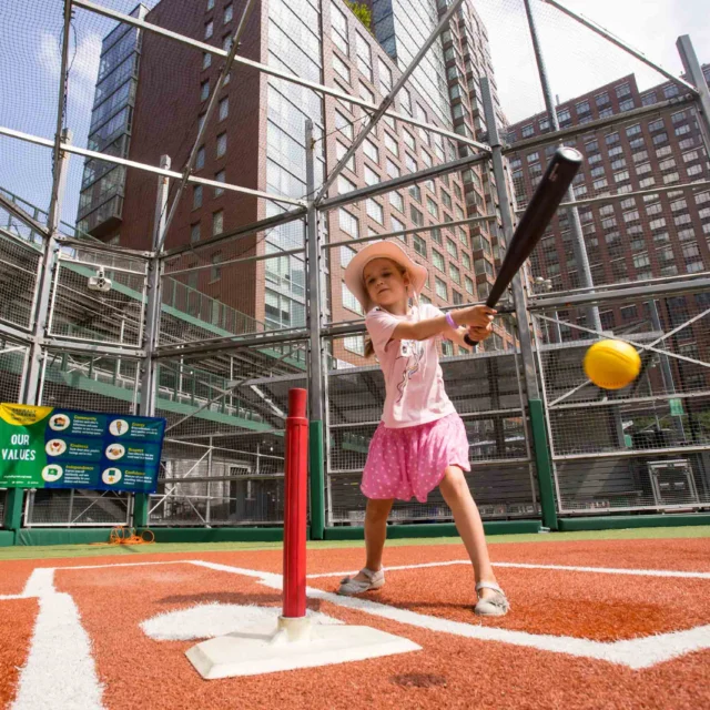A young girl in a pink outfit and hat swings a bat at a stationary ball on a tee in an outdoor urban baseball field.