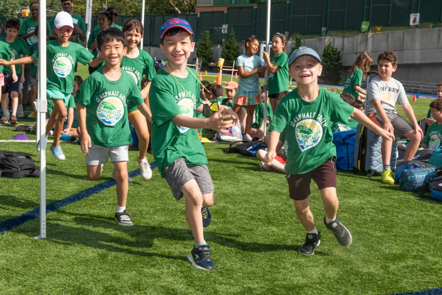 Group of kids wearing green shirts running and smiling energetically on a grass field during a sunny day, with other children in the background.