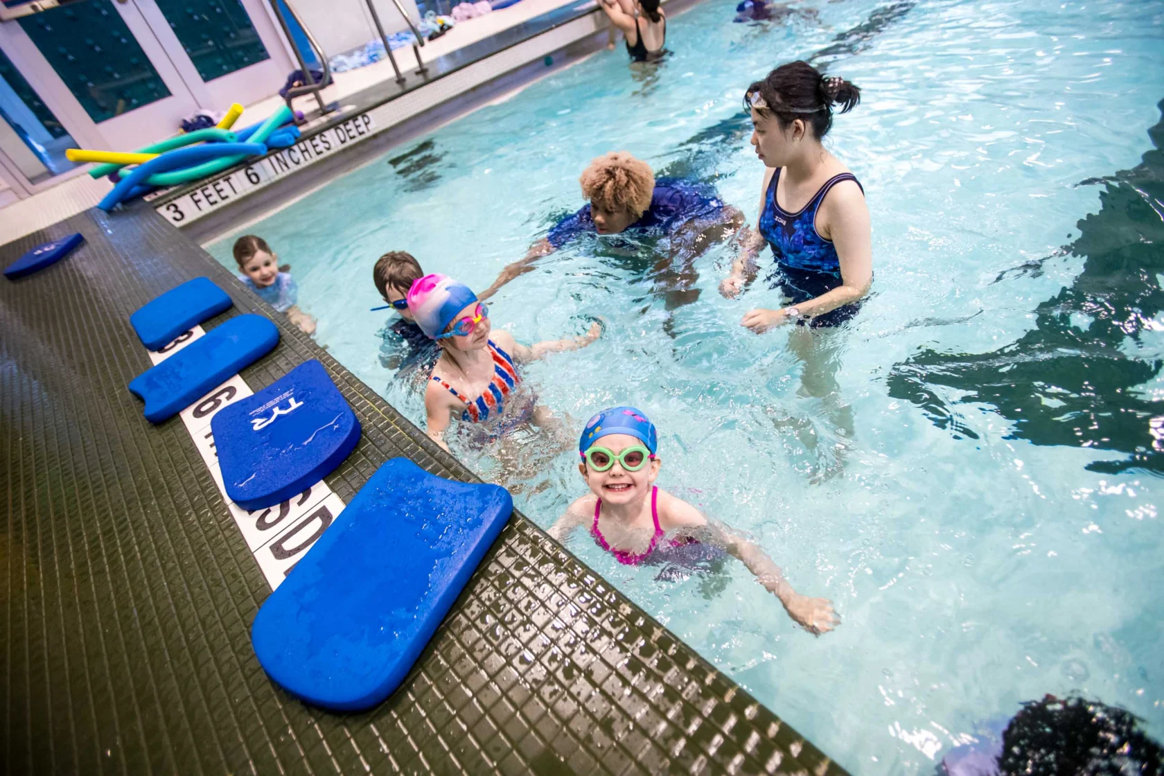 Children in a swimming pool during a lesson, guided by two instructors. Brightly colored swim gear is visible, and pool depth markings are shown.