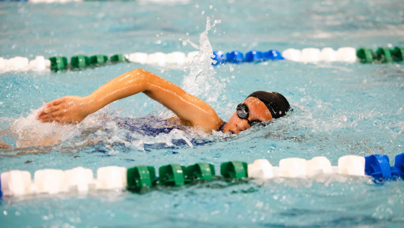 A swimmer in a black cap and goggles performs the freestyle stroke in a pool, surrounded by green and white lane dividers.