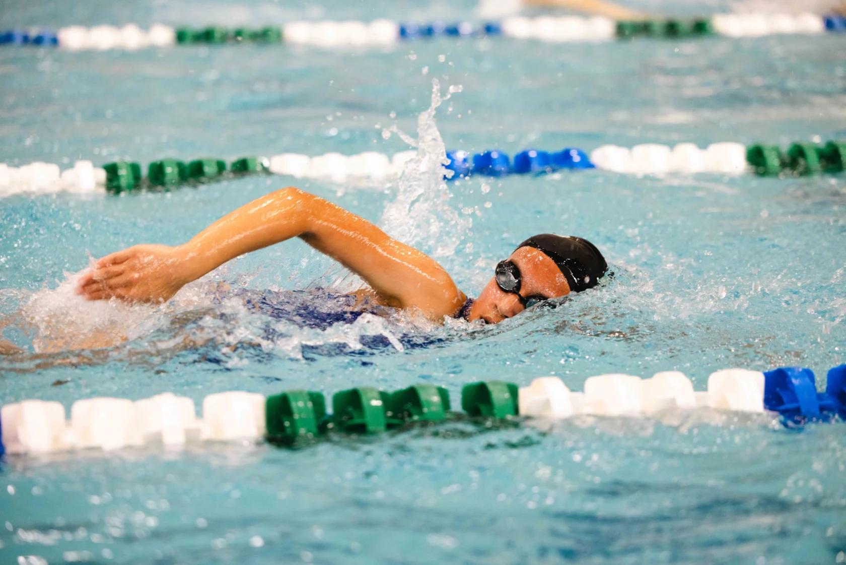 A swimmer in a black cap and goggles performs the freestyle stroke in a pool, surrounded by green and white lane dividers.