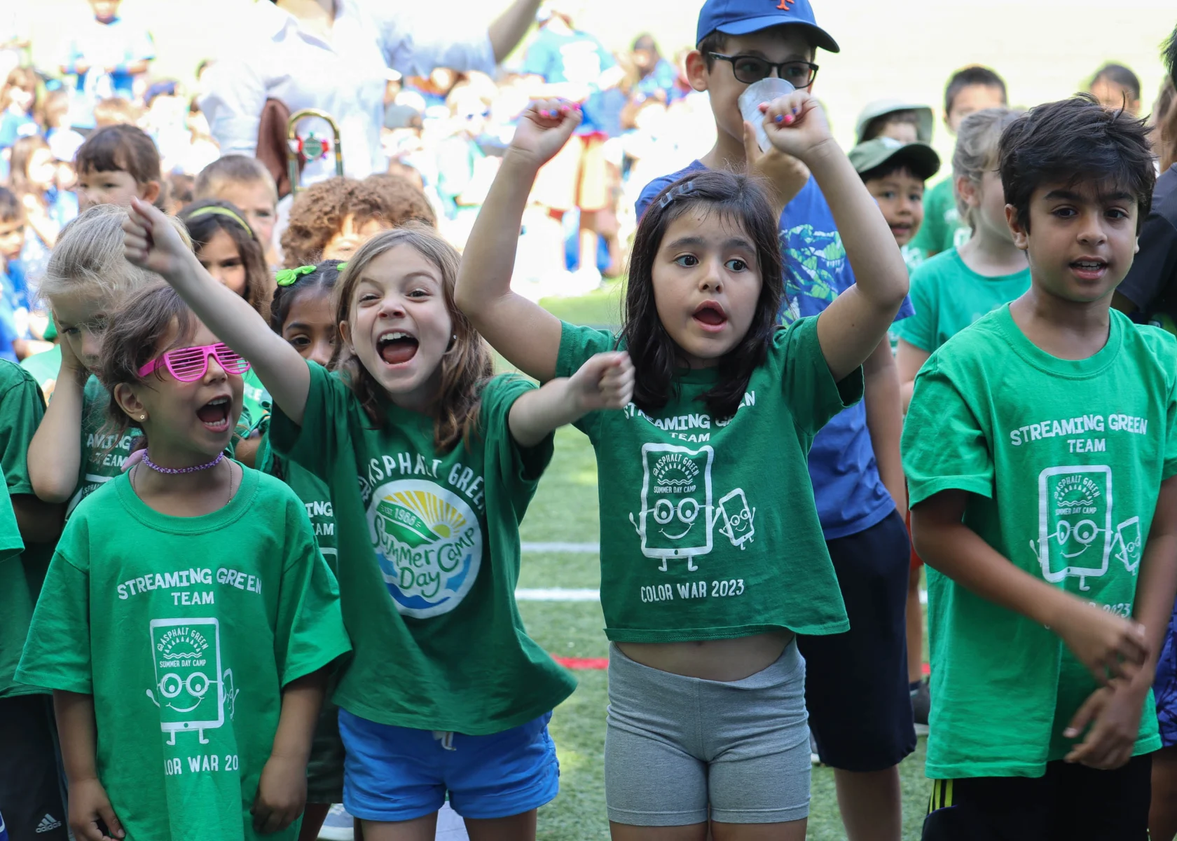 Children wearing green shirts cheer enthusiastically outdoors on a sunny day.